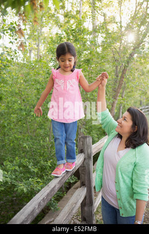 A young girl in a pink shirt and jeans, walking along a fence holding her mother's hand. Stock Photo