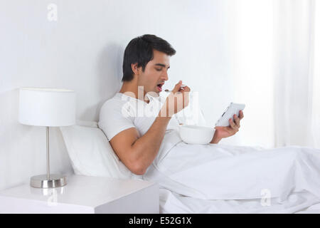 Side view of young man having breakfast while using digital tablet in bed Stock Photo