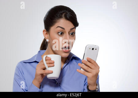 Shocked businesswoman with coffee mug reading text message on smart phone against gray background Stock Photo