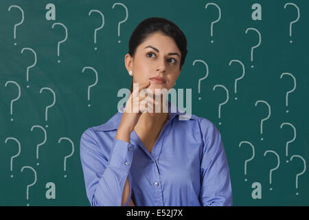 Confused young businesswoman standing against question marks on green board with question marks at office Stock Photo