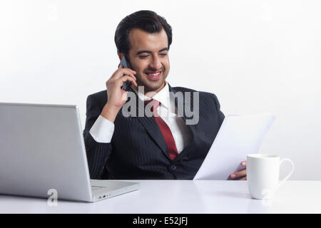 Happy businessman reading document while answering smart phone at desk over white background Stock Photo