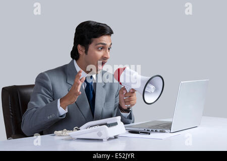 Aggressive businessman shouting through megaphone over laptop at desk against gray background Stock Photo