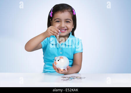 Portrait of happy girl putting coins in piggy bank against blue background Stock Photo