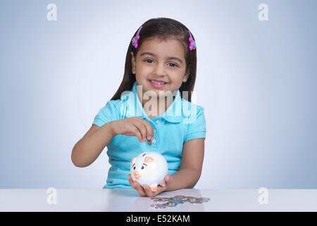 Portrait of cute girl putting coins in piggy bank against blue background Stock Photo