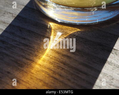 Refracted light through beer glass creating triangular highlight in the shadow on wood grain with reflection of blue sky in base Stock Photo