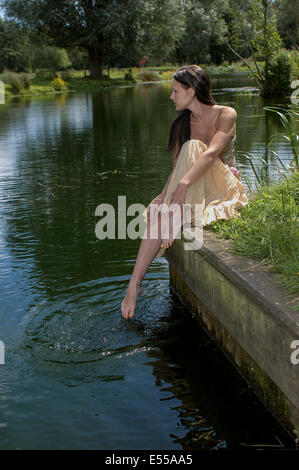 Young woman in summer dress sat on river bank with toes in the water Stock Photo