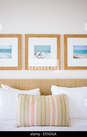 Hotel bed with striped pillow, white linens, and triptych of ocean images in wood frames. Stock Photo