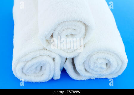 Three large white cotton bath cloths rolled up individually and piled together on a blue background. A few white cotton bath towels rolled up on blue. Stock Photo
