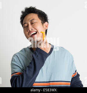 Young southeast Asian guy shaving his beard in a morning, on plain background Stock Photo