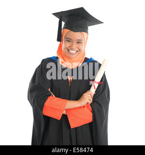 Portrait of smiling Asian female Muslim student in graduate gown showing graduation diploma standing isolated on white backgroun Stock Photo