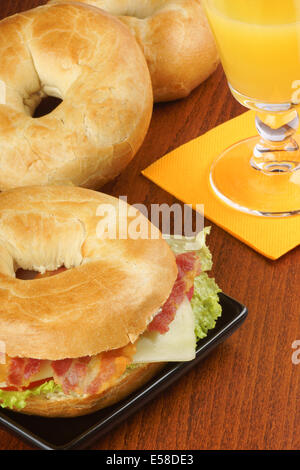Bagel with bacon, cheese and lettuce served on a black dish and a glass of orange juice over a wooden background. Stock Photo