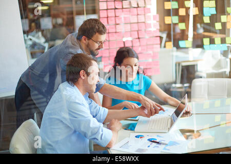 Group of three successful business partners in casual looking at laptop screen while two young men pointing at it at meeting in Stock Photo