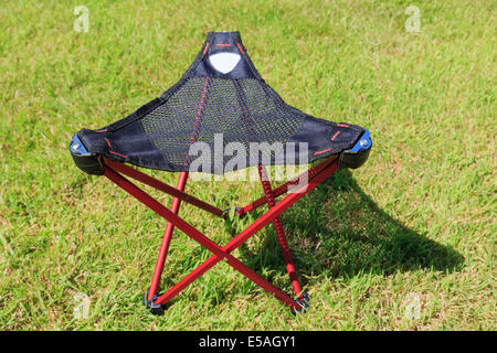 Folding portable three-legged tripod camping stool with a triangular mesh seat on grass in sunshine Stock Photo