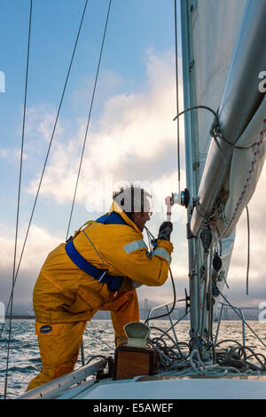 Sailor in yellow foul weather gear securing the halyard after raising the mainsail heading into cloudy weather in his sailboat Stock Photo