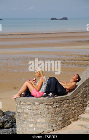couple relaxing in the sun with the remnants of Mulberry Harbour on the beach at Arromanches Normandy France in July Stock Photo