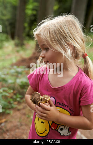 Portrait of happy child blond girl picking and carrying full hands of mushrooms outside in forest Stock Photo