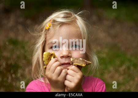 Portrait of happy child blond girl picking and carrying full hands of mushrooms outside in forest Stock Photo