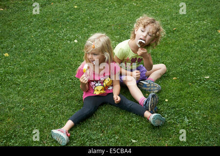 Children blond girls eating ice cream cones sitting on grass lawn, summertime Stock Photo