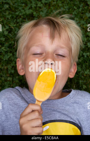 Blond child 6-8 year old boy holds, lickling and eating yellow ice cream summertime laying on green grass lawn background Stock Photo