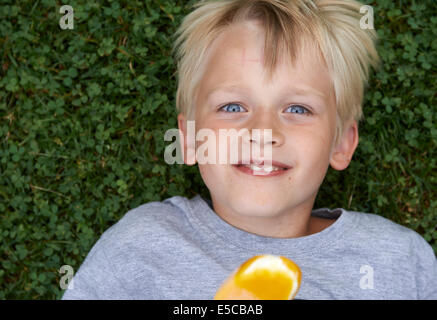 Blond child 6-8 year old boy holds, lickling and eating yellow ice cream summertime laying on green grass lawn background Stock Photo