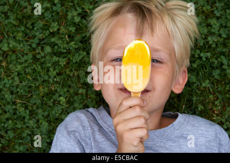 Blond child 6-8 year old boy holds, lickling and eating yellow ice cream summertime laying on green grass lawn background Stock Photo