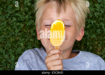 Blond child 6-8 year old boy holds, lickling and eating yellow ice cream summertime laying on green grass lawn background Stock Photo