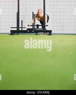 Strong young woman pushing the prowler on artificial grass turf. Fit female using prowler exercise equipment at the crossfit gym Stock Photo