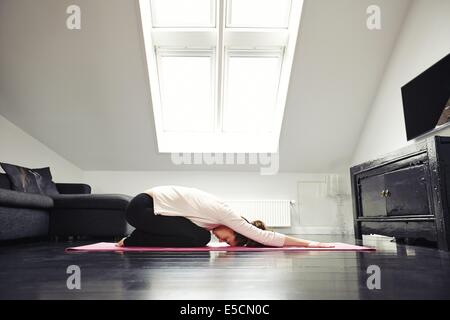 Side view of young caucasian woman exercising yoga in her living room. Female doing relaxation exercise on mat at home. Stock Photo