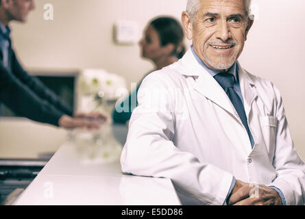 Smiling senior male doctor smiling at hospital reception desk, female patient speaking with young doctor on background. Stock Photo