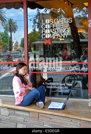 woman chats on phone and works on laptop while sitting on coffee shop bench Stock Photo