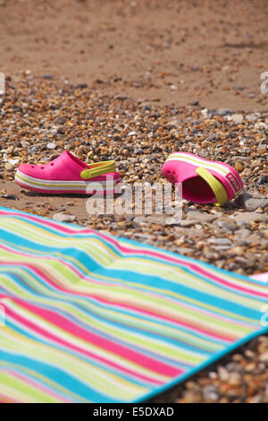 pink crocs discarded on beach next to stripey beach towel Stock Photo