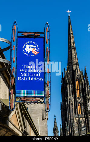 A Tartan Weaving Mill and Exhibition sign hangs on a shop on the Royal Mile with the Gothic spire of the Hub behind it.
