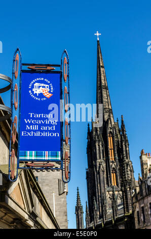 A Tartan Weaving Mill and Exhibition sign hangs on a shop on the Royal Mile with the Gothic spire of the Hub behind it.