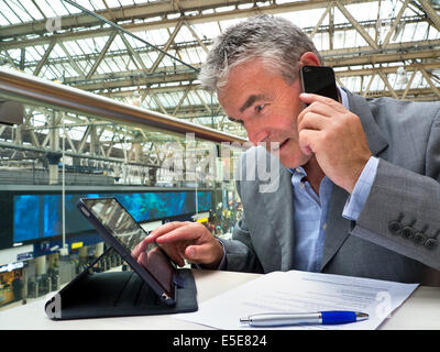 Mature businessman seated at cafe table on railway concourse looking at his tablet computer screen and using his smartphone Stock Photo