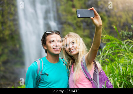 Couple having fun together outdoors. Taking self portrait with camera phone after hiking to incredible waterfall in Hawaii. Stock Photo