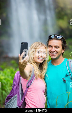Couple having fun together outdoors. Taking self portrait with camera phone after hiking to incredible waterfall in Hawaii. Stock Photo