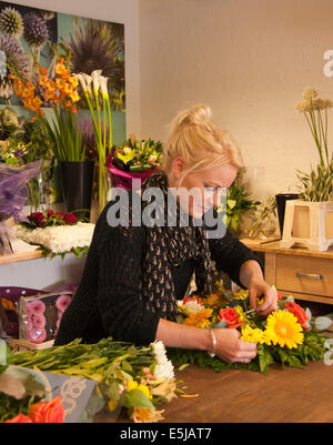 Florist arranging a wreath in a florist's shop with bunches of flowers and a flower poster in the background. Stock Photo