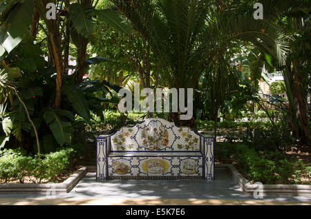 An ornate ceramic tiled bench sits in the shade surrounded by lush green botanical plants in Parque de Alameda park Marbella Spain. Stock Photo