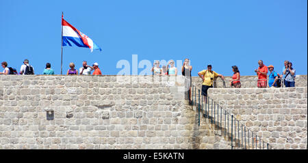 Tourists walking old city walls of Dubrovnik with Croatian national flag fluttering in breeze on hot summer blue sky day Croatia Dalmatia Adriatic Stock Photo