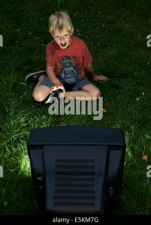 Child Blond Boy Watching Television in Yard, outside in green grass lawn, emotional, facial expression, grimace, grinning Stock Photo
