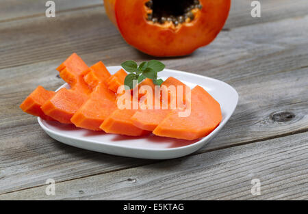 Closeup view of sliced papaya fruit, on white plate, with mint leaf and half piece in background on rustic wood Stock Photo