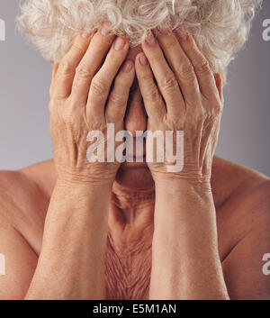 Close-up portrait of a sad looking senior woman with her head in her hands against grey background. Upset old woman covering her Stock Photo