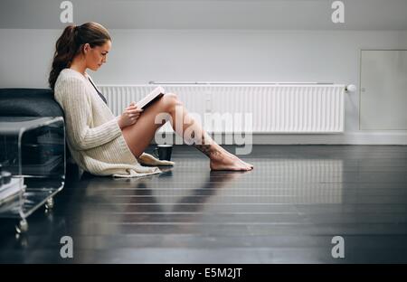 Side view of young woman relaxing on floor reading book. Pretty young lady sitting on floor at home reading a novel. Stock Photo