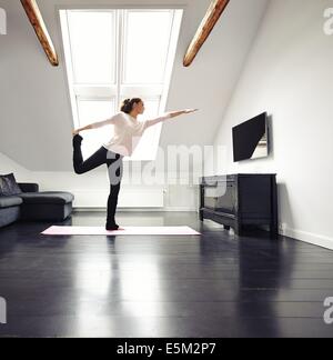 Beautiful woman practicing yoga exercises in living room. Fit female standing on one leg exercising at home. Stock Photo