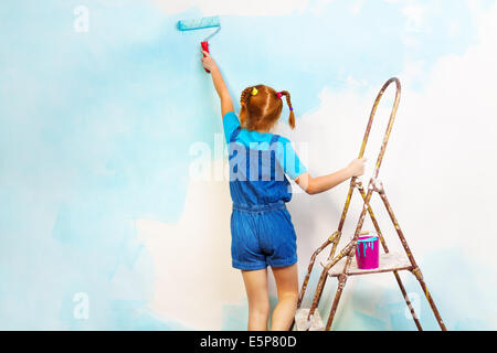 Little girl in blue paints the wall on a ladder Stock Photo