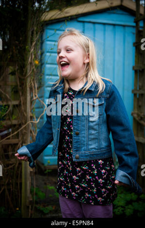 happy Blonde girl of 8 9 10 in blue denim jacket standing in front of blue shed Stock Photo