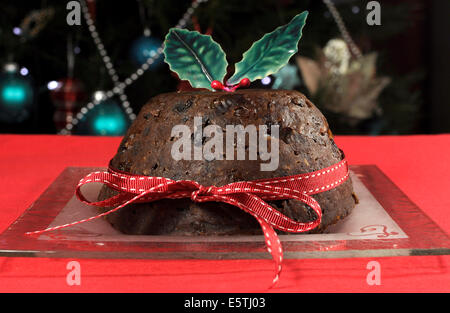 Beautiful Christmas table setting in front of Christmas Tree, featuring a classic plum pudding with holly on red tablecloth. Stock Photo