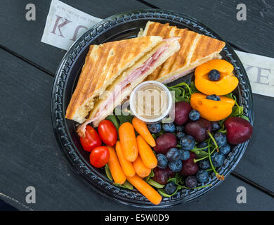 A lunch plate filled colorful fruits and vegetables and a toasted ham and cheese sandwich sits on a picnic table. Stock Photo