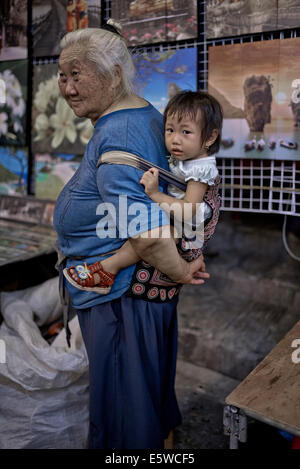 Grandmother carrying infant granddaughter in a back sling harness. Thailand S. E. Asia, Asian grandma carrying child Stock Photo