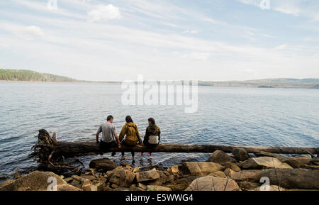 Three hiking friends sitting in a row on fallen tree at lakeside Stock Photo